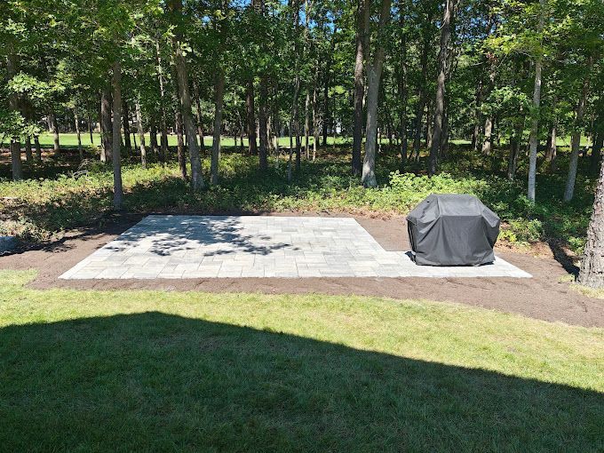 A new patio area built by stone works masonry inc. with a grill, surrounded by grass and trees in a backyard setting