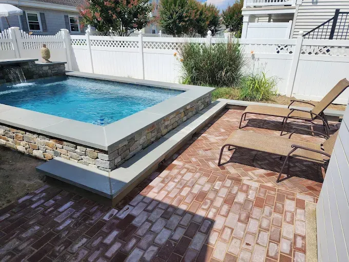 A swimming pool is surrounded by brick patio and chairs in a backyard built by stone works masonry inc.