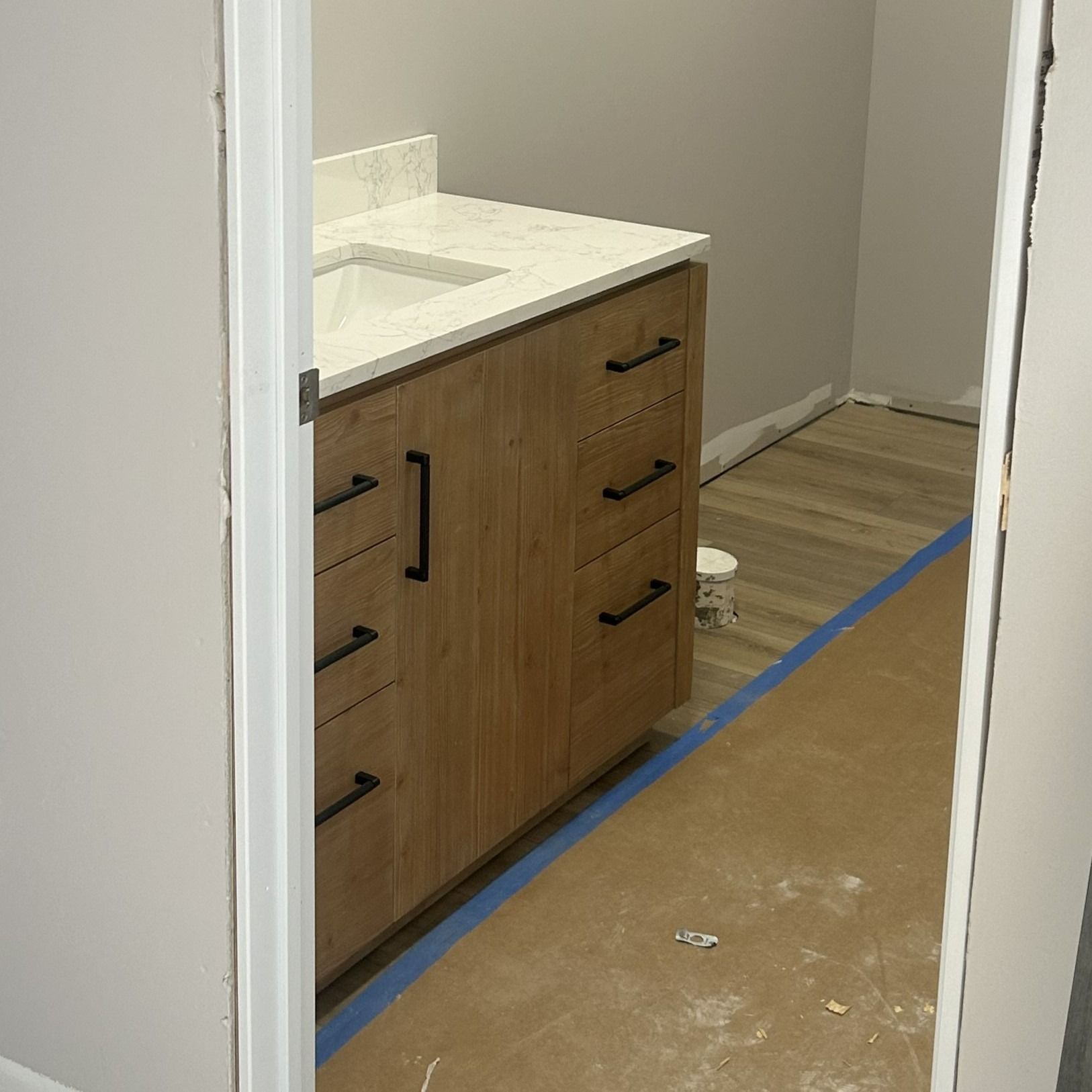 Bathroom with light wood vanity, white countertop, and gray walls, viewed through a doorway.