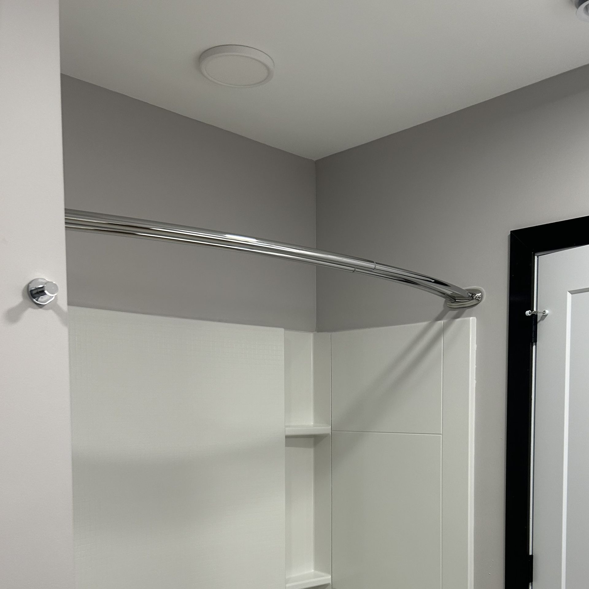 Corner of a white shower stall with a curved chrome shower rod, gray walls, and a recessed ceiling light.