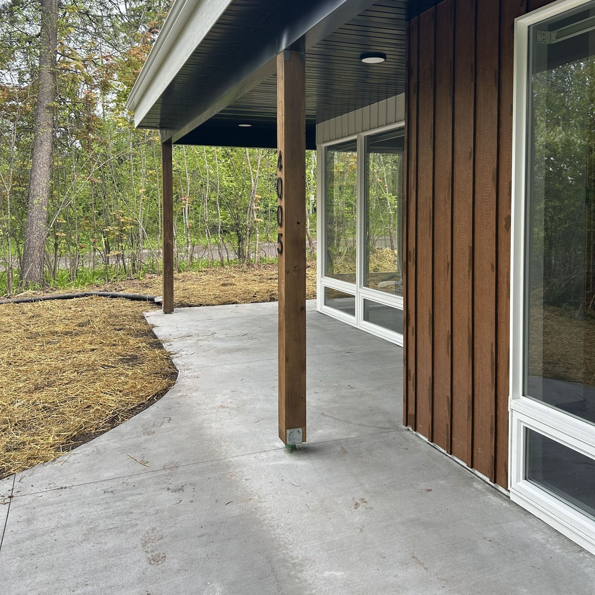 Exterior of a modern home with concrete porch, brown siding, wooden supports, and large windows.