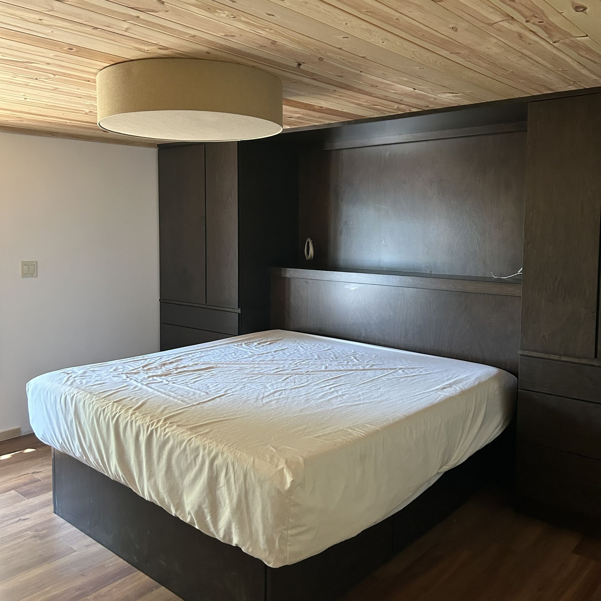 Bedroom with a large bed, built-in dark cabinets, wood ceiling and floor, and round light fixture.