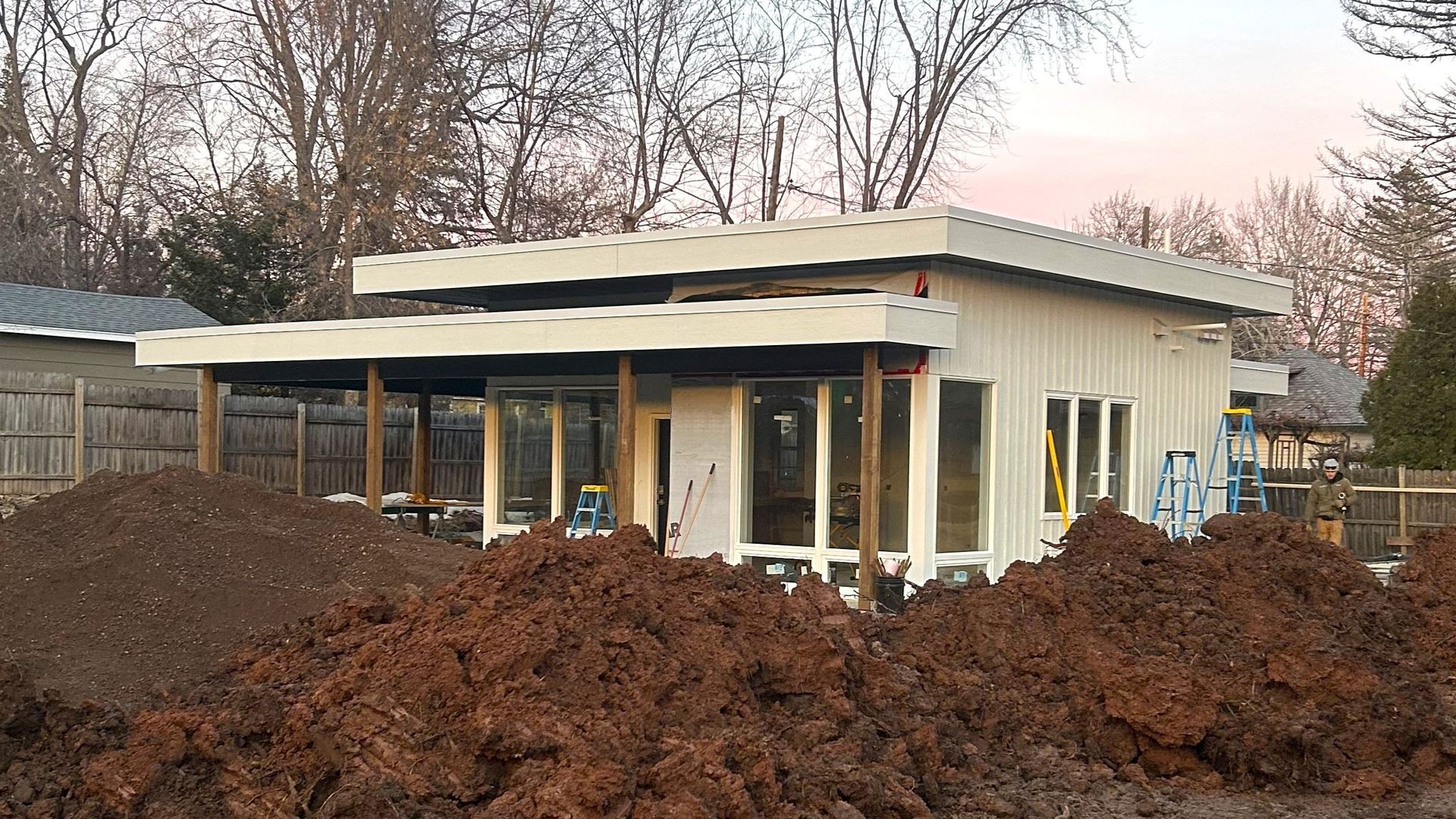 Modern, single-story house under construction, with large windows, covered porch, and piles of dirt in front.
