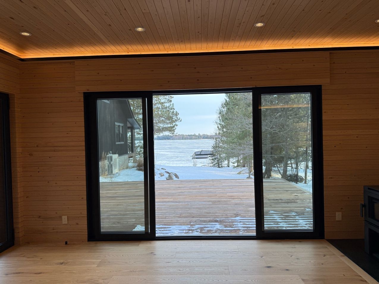 Wooden-paneled room with large black-framed windows overlooking a snowy lake and trees.