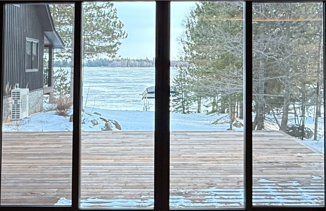 View of a snowy lake and trees from a wooden deck, through a window. A cabin is visible to the left.