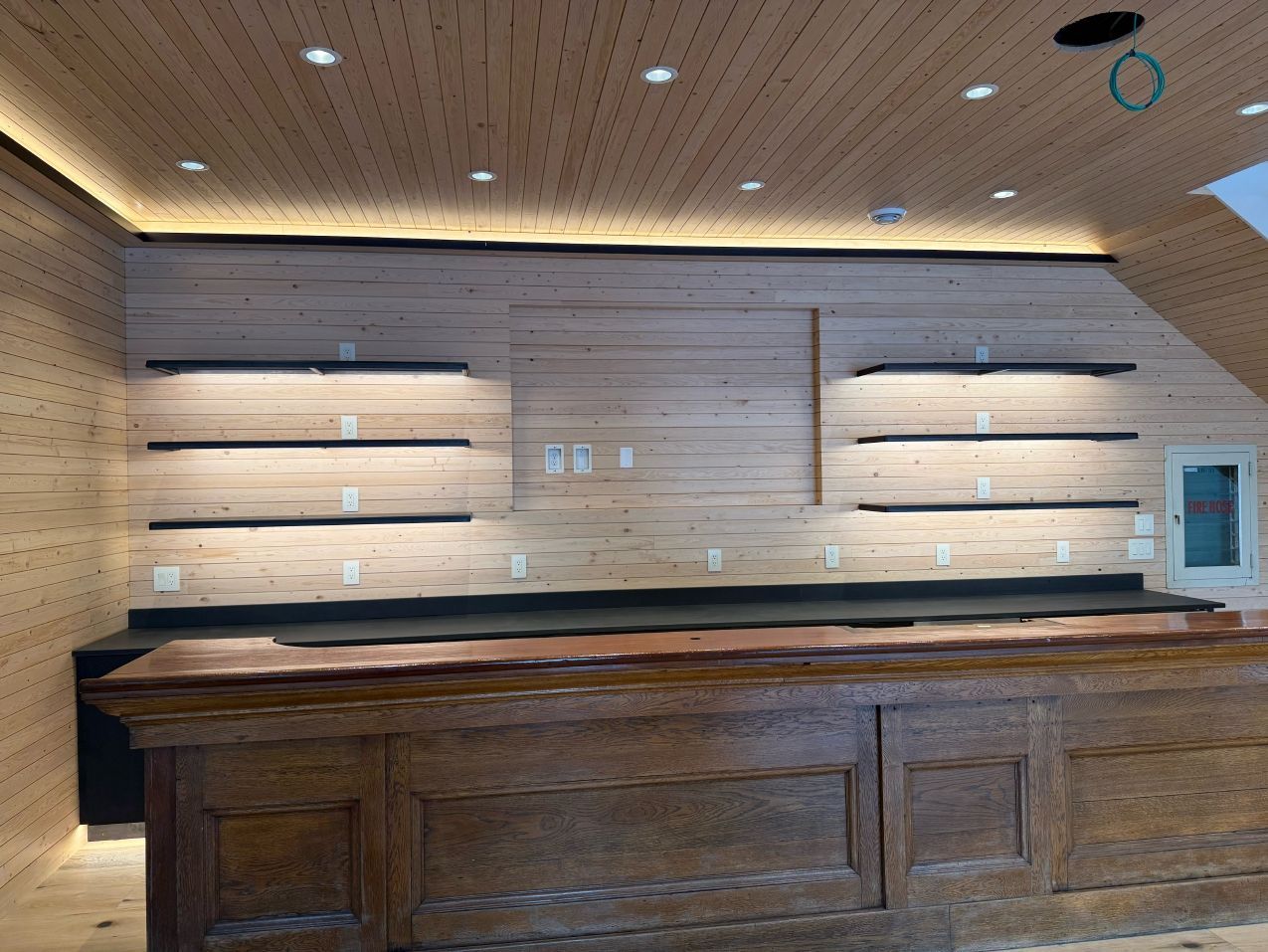 Bar interior with wooden walls, bar, and shelves; recessed lighting.