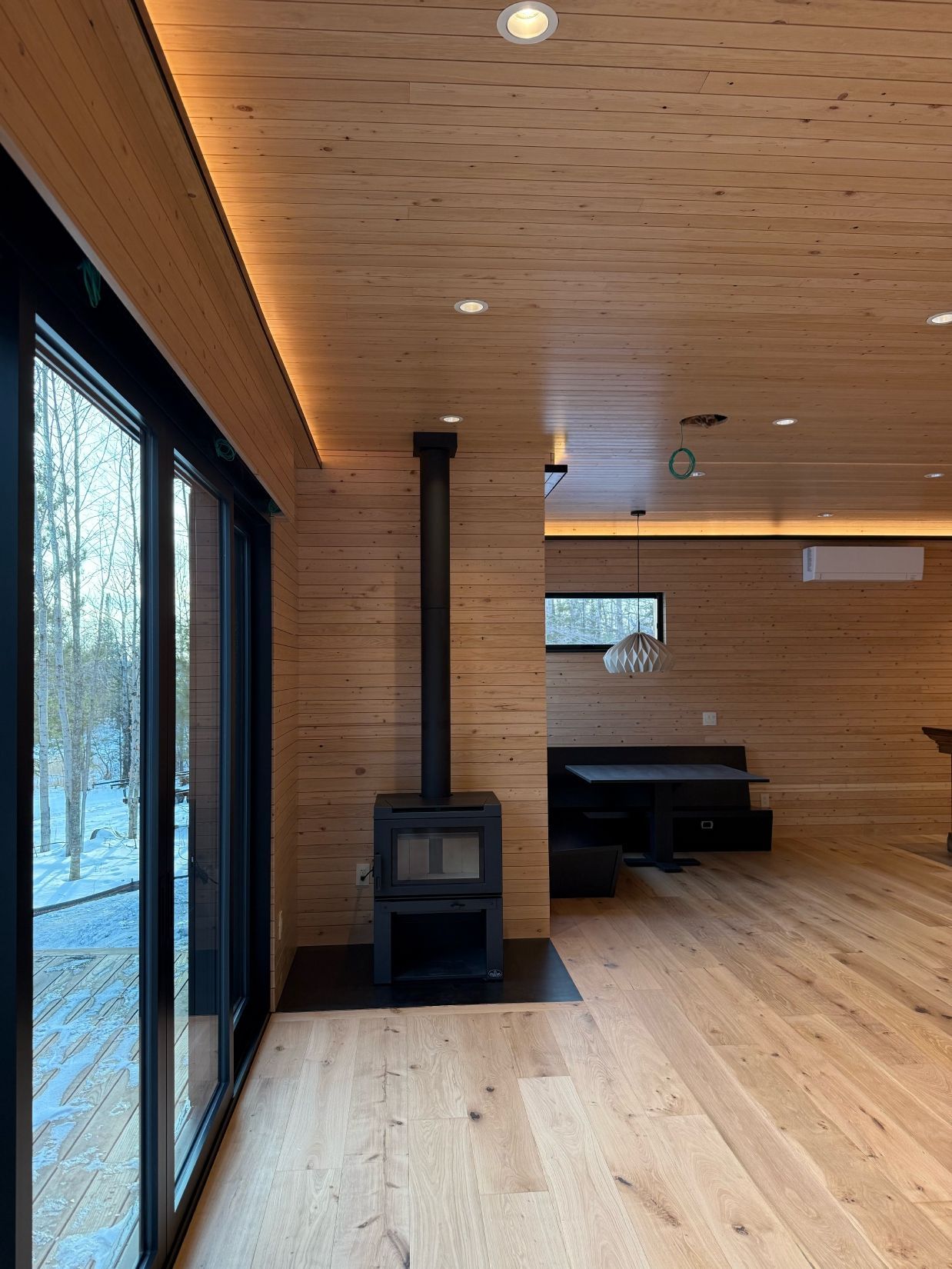 Interior of a modern cabin, featuring a wood-burning stove, large windows, and wooden walls/ceiling.