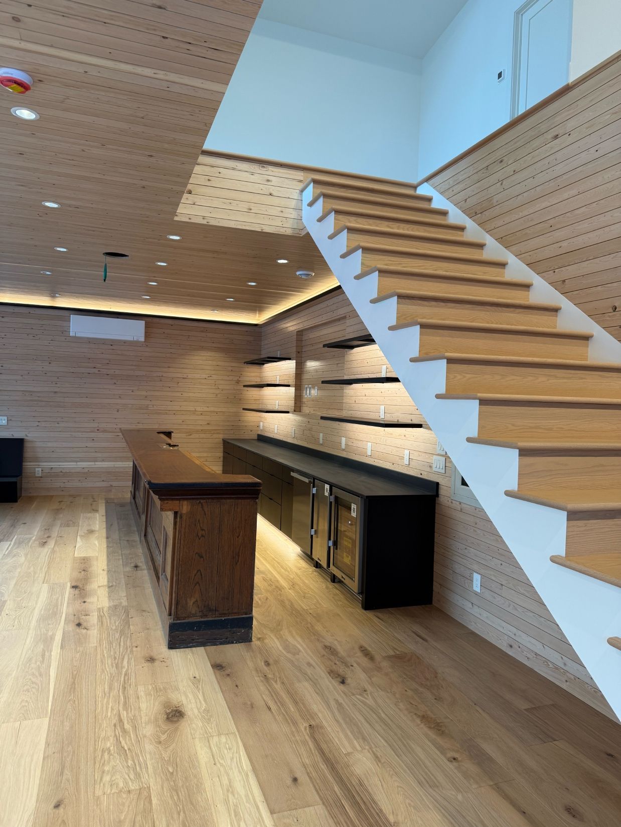 Wooden bar and staircase in a room with light wood floors and walls, recessed lighting.