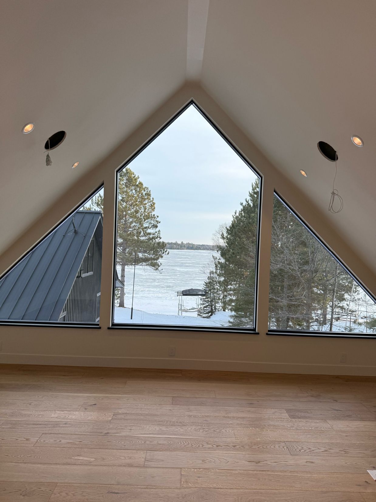 Triangular window framing a snowy lake view, with trees and a dark roof visible. Interior, bright ceiling, wood floor.