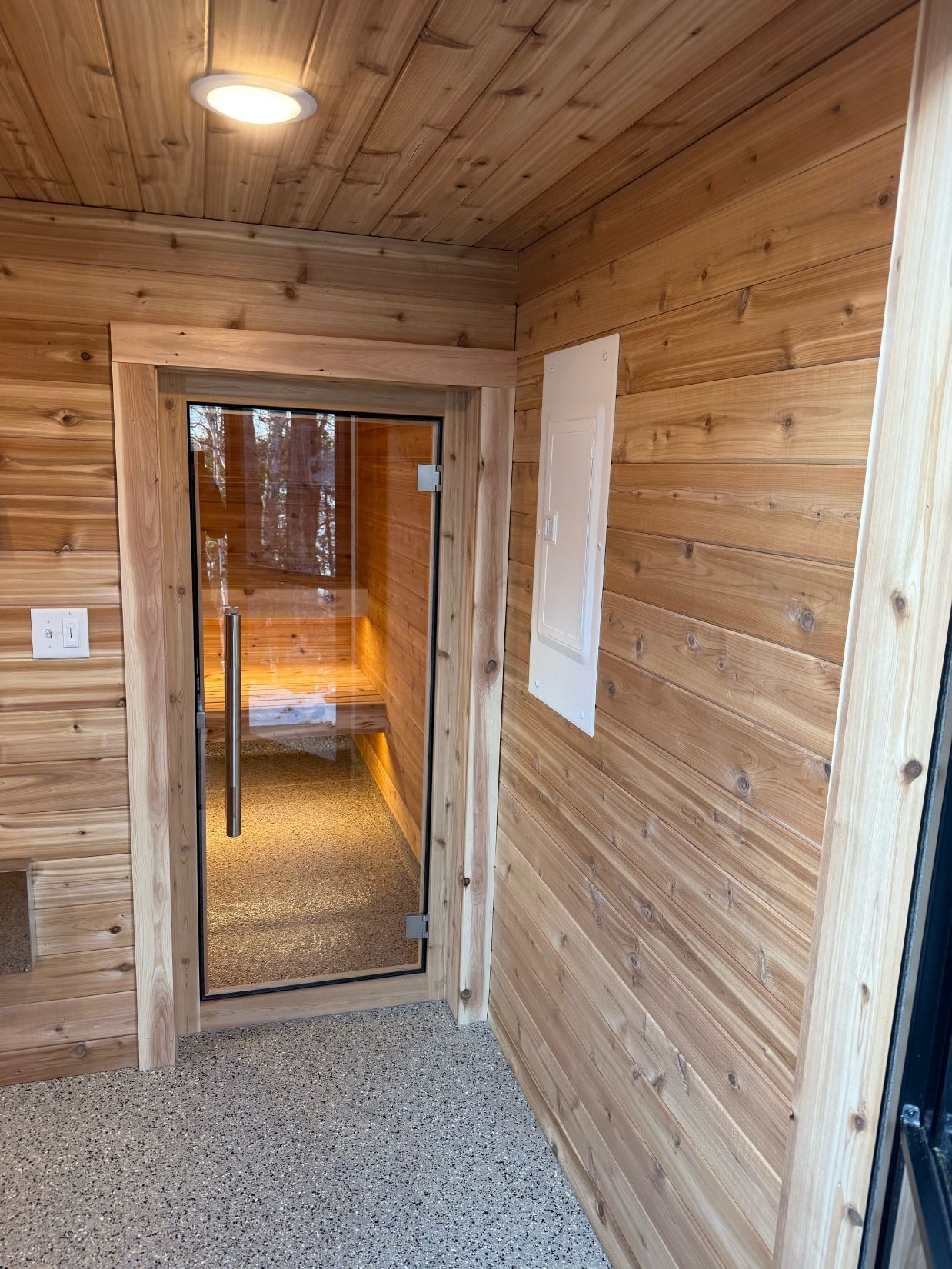 Wooden sauna interior with glass door, light fixtures, and gravel floor.