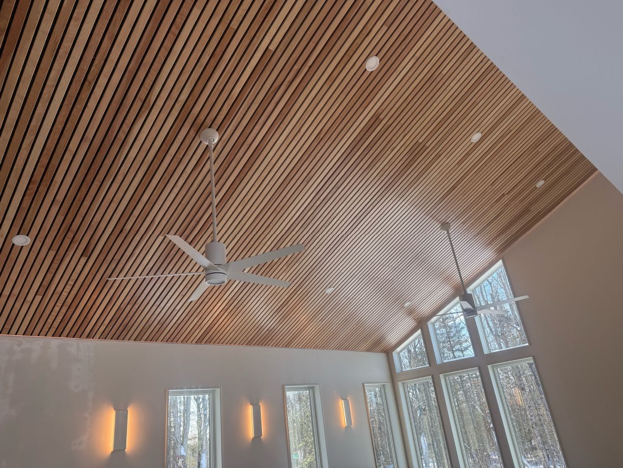 Wooden slatted ceiling with two ceiling fans, recessed lights, and large windows.