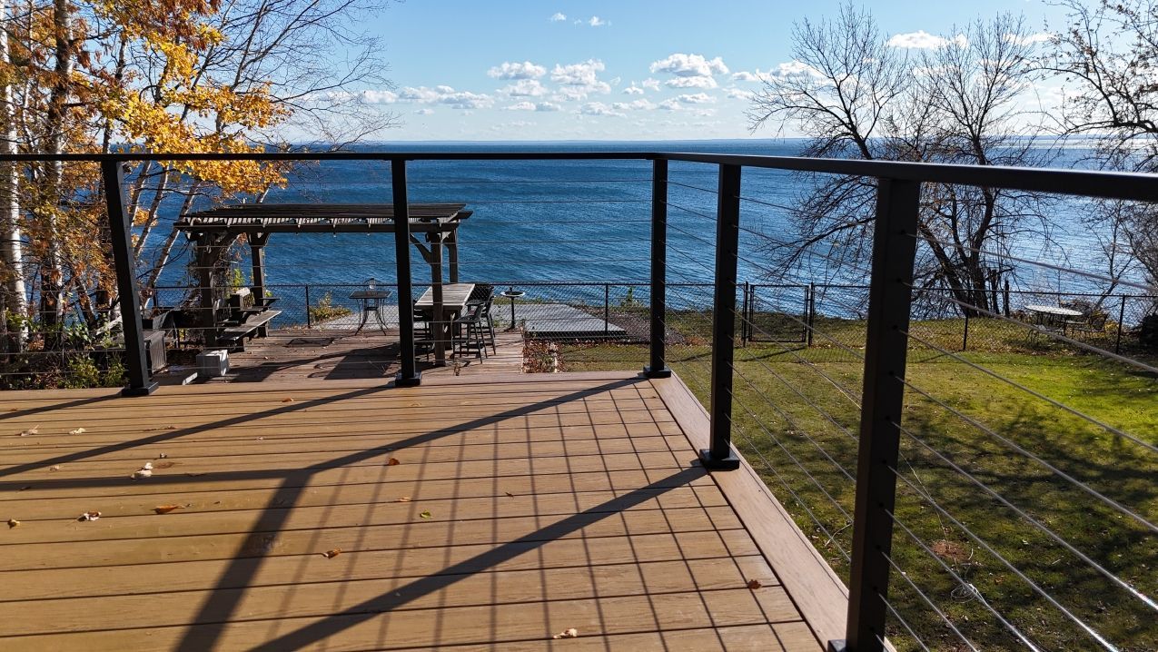 Deck overlooking blue water, gazebo, and trees under a clear sky. Black railing.