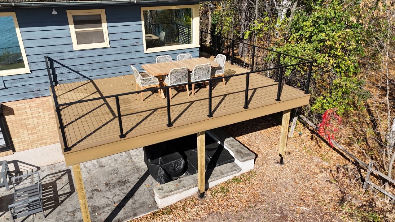 Deck with table, chairs, and black railing next to blue house and trees.