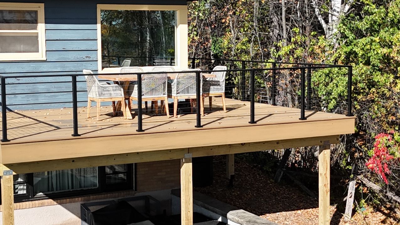 Wooden deck with black railings, table, chairs, overlooking a wooded area.