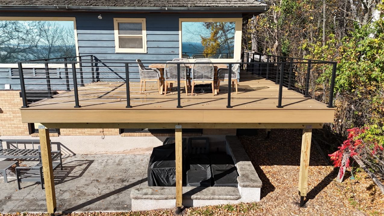 Deck with black railings, tan deck boards, blue house with reflective windows, chairs, and surrounding trees.