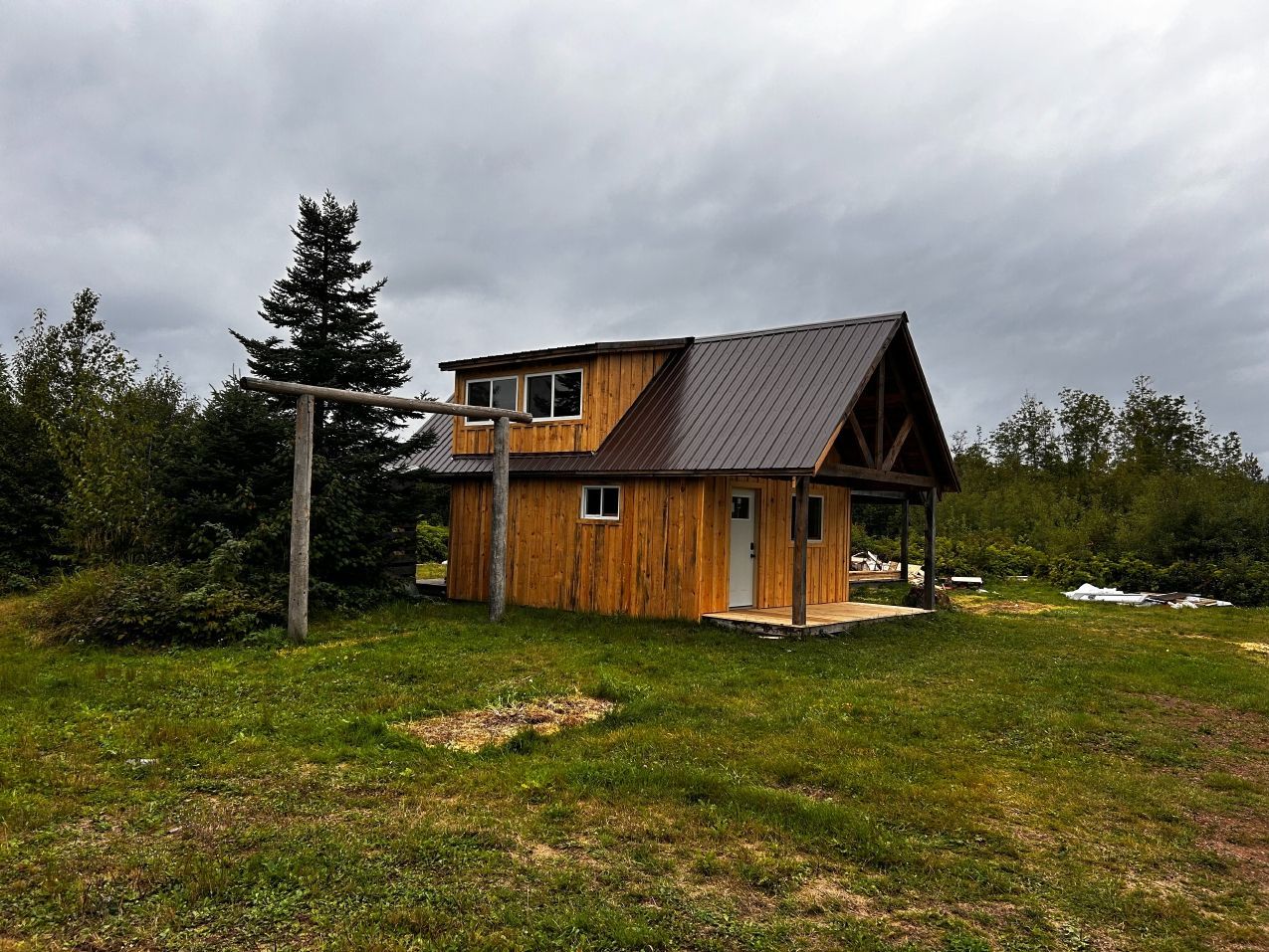 Wooden cabin with brown roof and porch, on a grassy field under a cloudy sky.