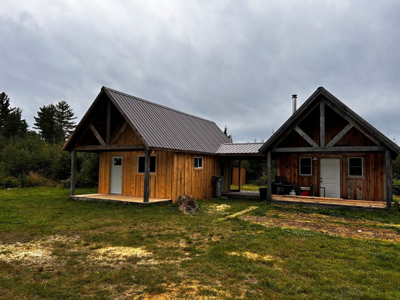 Two rustic wooden cabins with porch and connecting covered walkway, set in a grassy clearing under a cloudy sky.