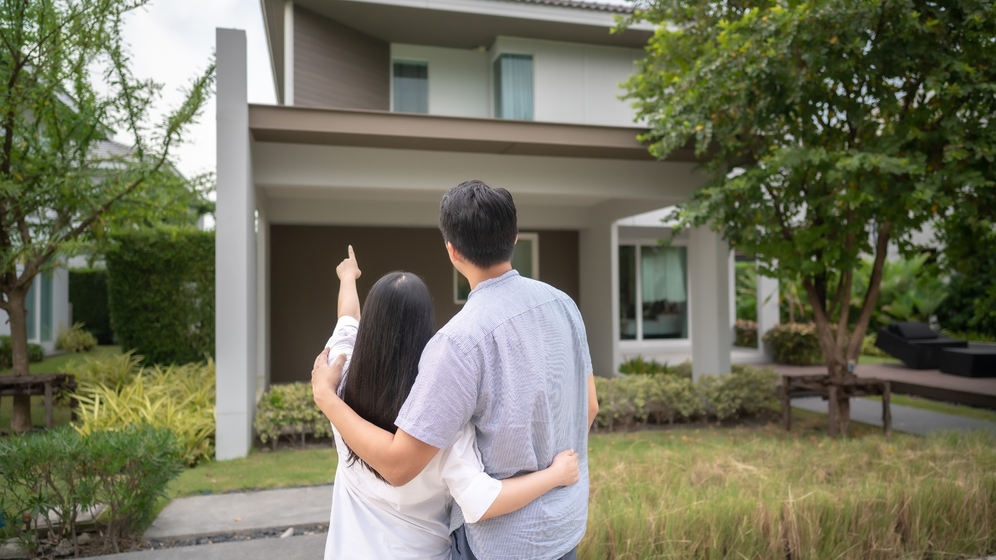 Couple standing in front of a house, woman pointing upwards, green yard, sunny day.