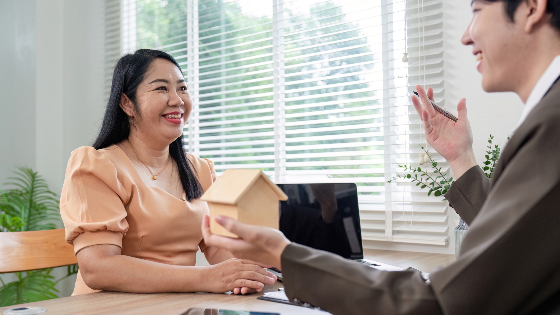 Woman and realtor in an office, smiling. Realtor gestures towards a small house model on the desk.