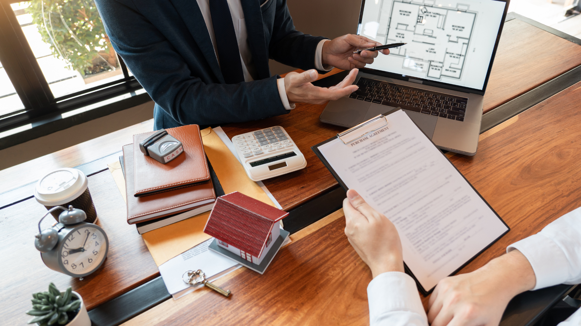 Real estate agent pointing at a blueprint on a laptop, discussing a document with a client.