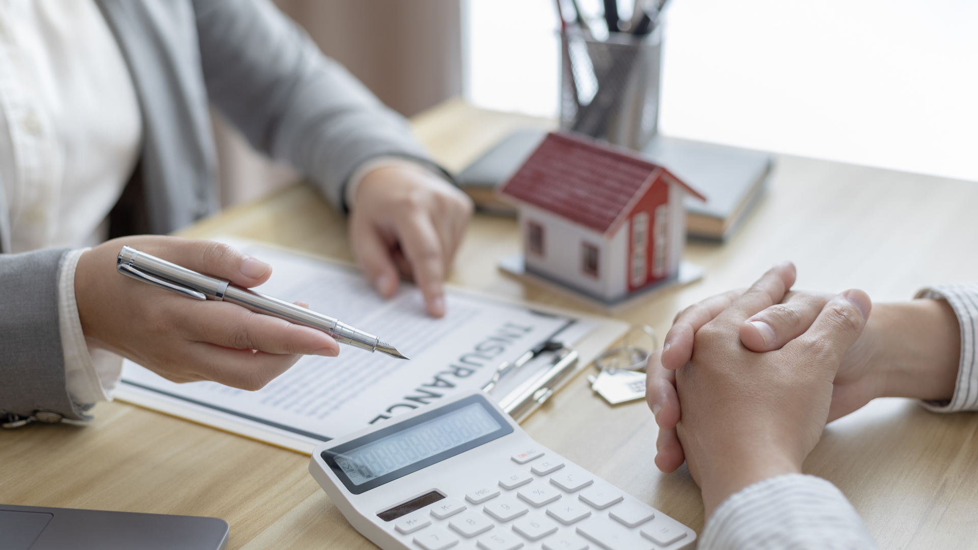 Person pointing at insurance document with pen, discussing with another person. Model house and calculator on the table.