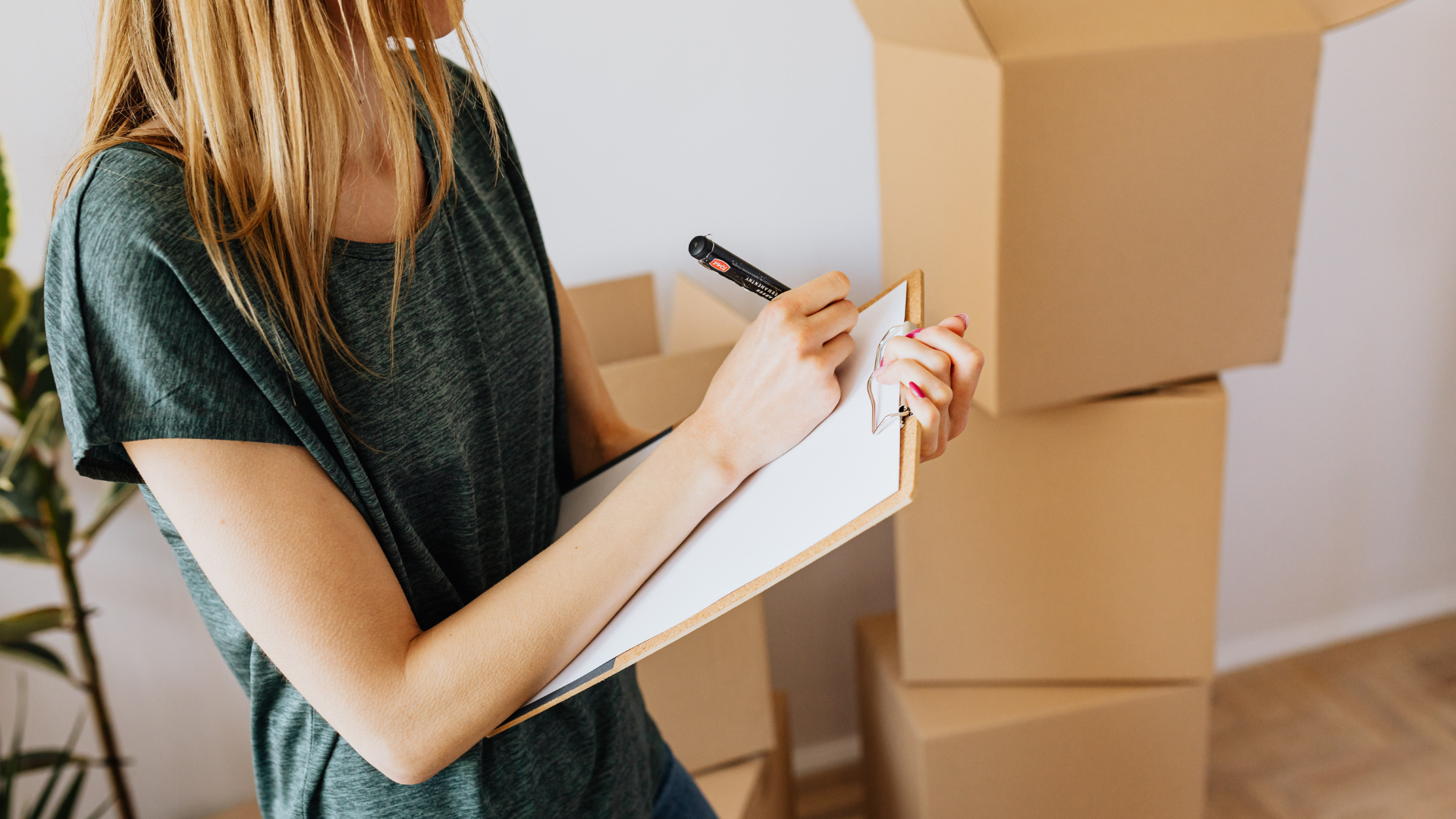 Woman in green shirt writing on clipboard next to cardboard boxes, indoors.