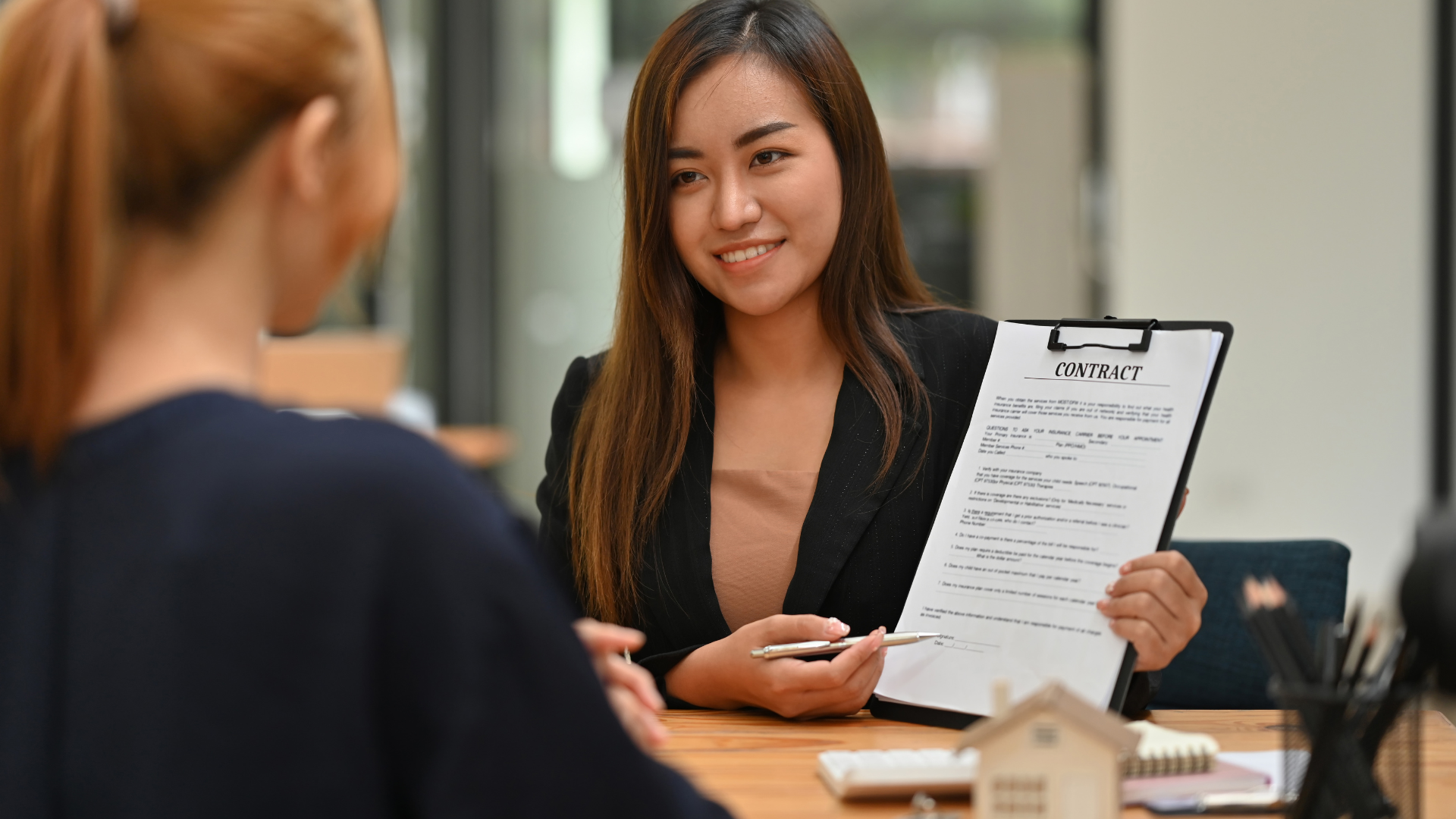 Real estate agent showing client a house model and paperwork at a desk.