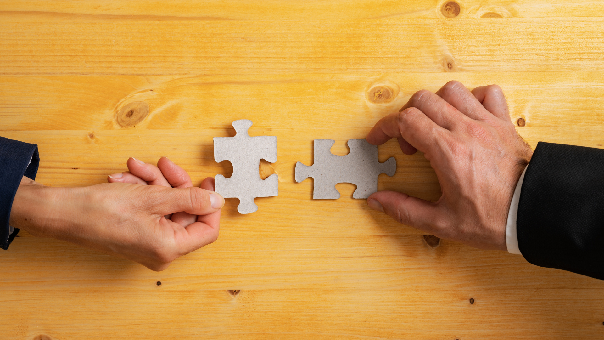 Two hands connecting puzzle pieces on a wooden table.