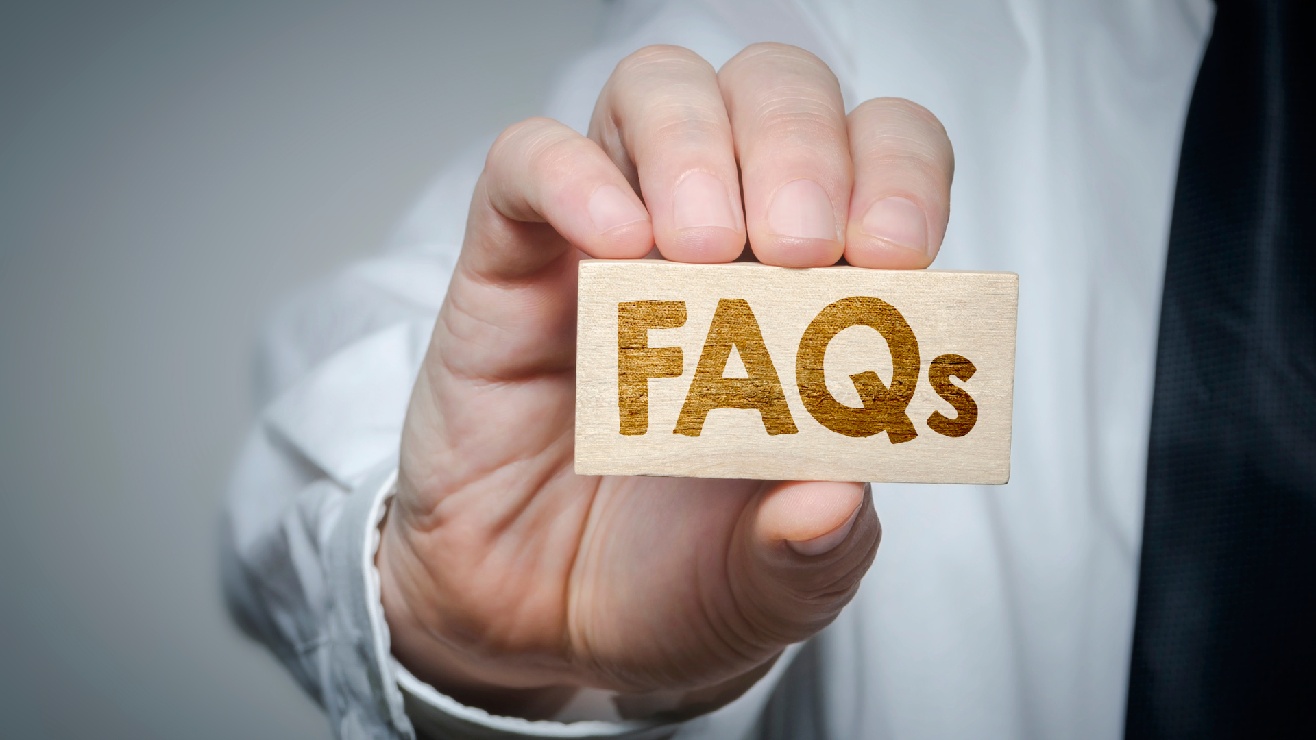 Person holding a small wooden sign that reads “FAQs” in brown letters.
