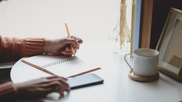 Person writing with a pencil in a notebook by a window with a coffee mug and phone.