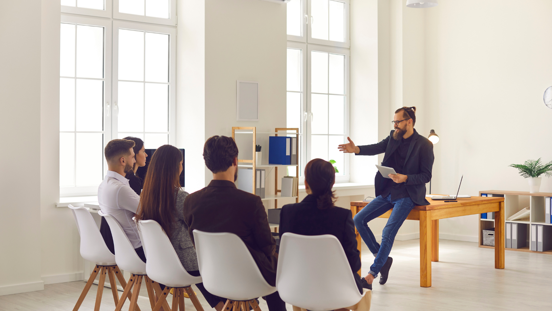 A man gestures while addressing a seated group in a bright office; sunlight streams through windows.