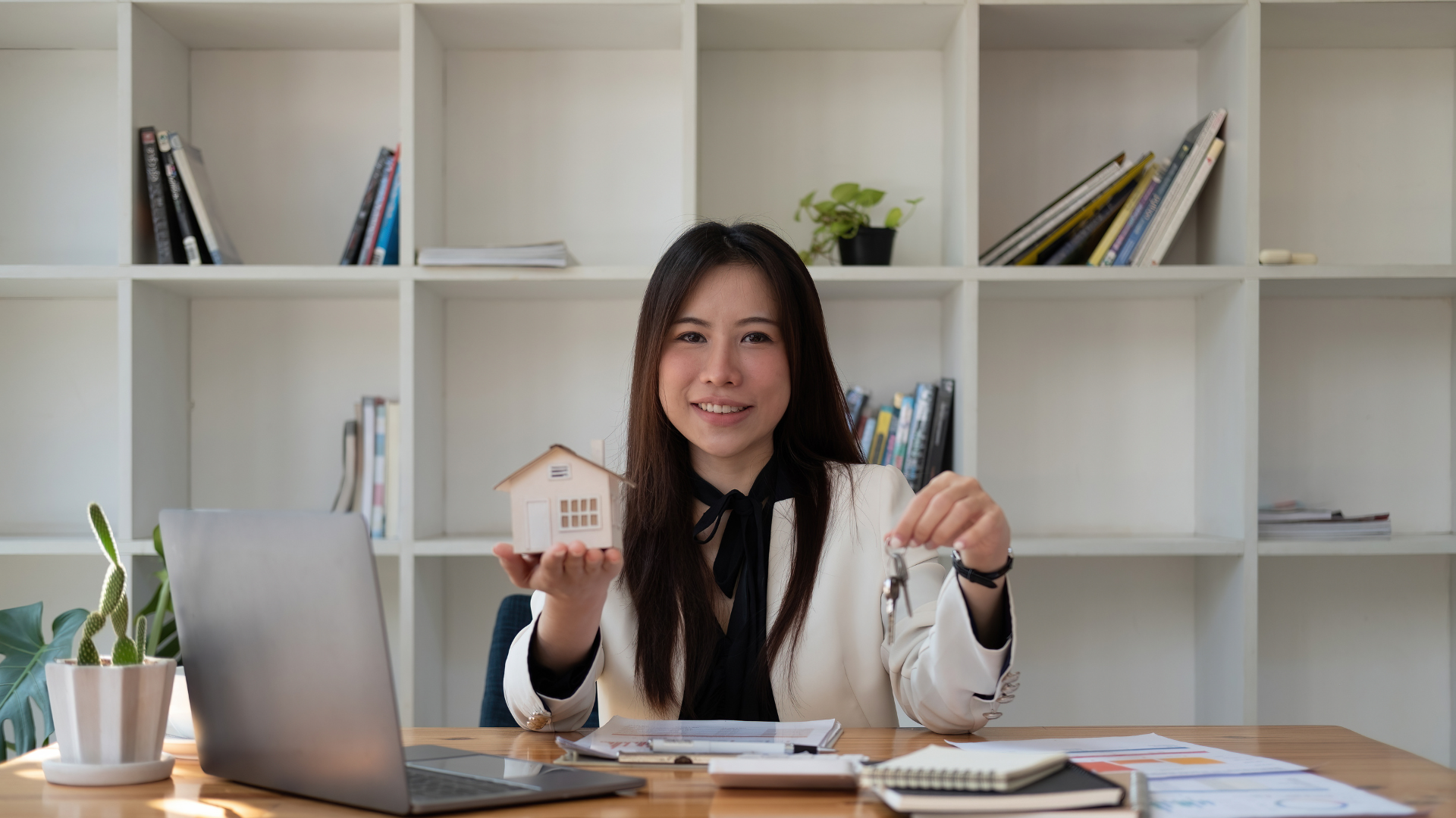 Woman real estate agent holding a house model and keys, smiling at a desk with a laptop.
