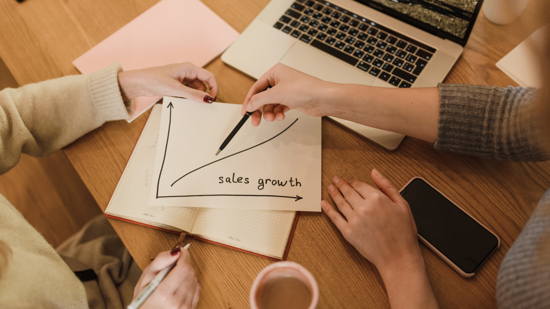Two people analyzing sales growth chart with laptop and phone on wooden table.