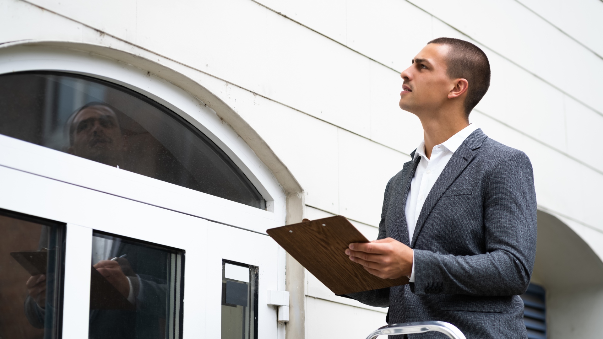 Man in a suit inspects a building, holding a clipboard and looking up at an arched window.