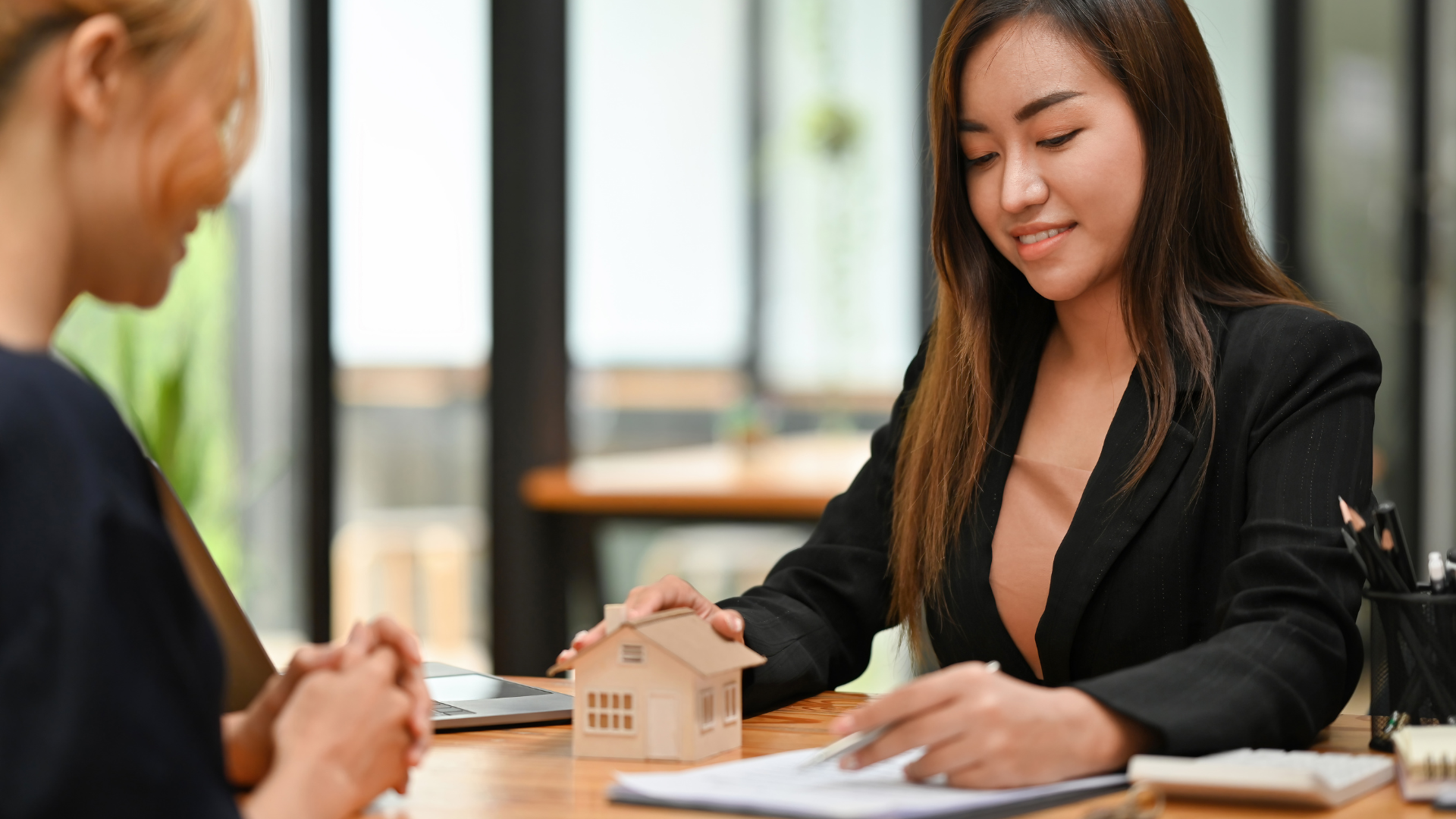 Real estate agent showing client a house model and paperwork at a desk.