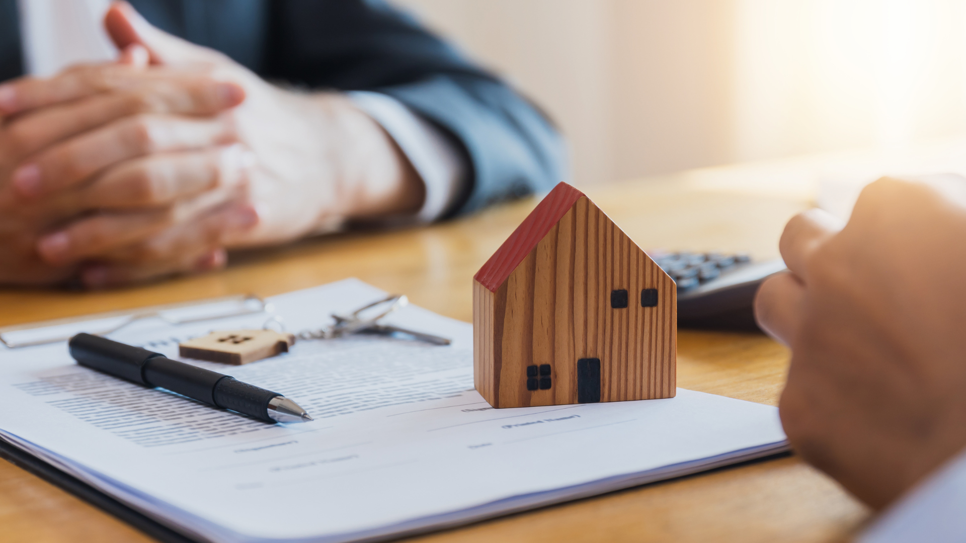 Wooden house model, keys, pen, and documents on a desk with people, representing a real estate deal.