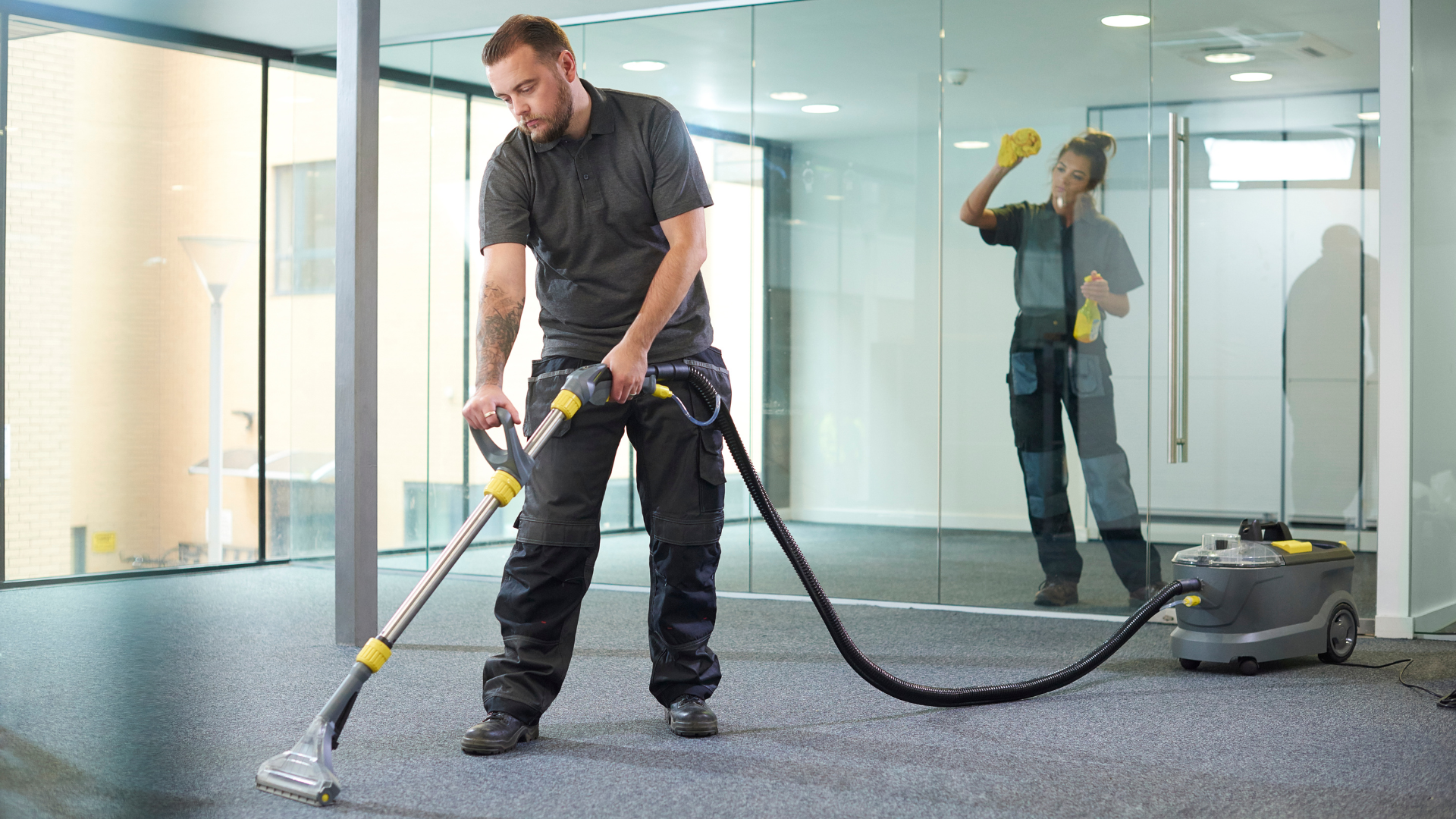 Two cleaners in an office cleaning carpet and glass. One uses a carpet cleaner, the other wipes a window.