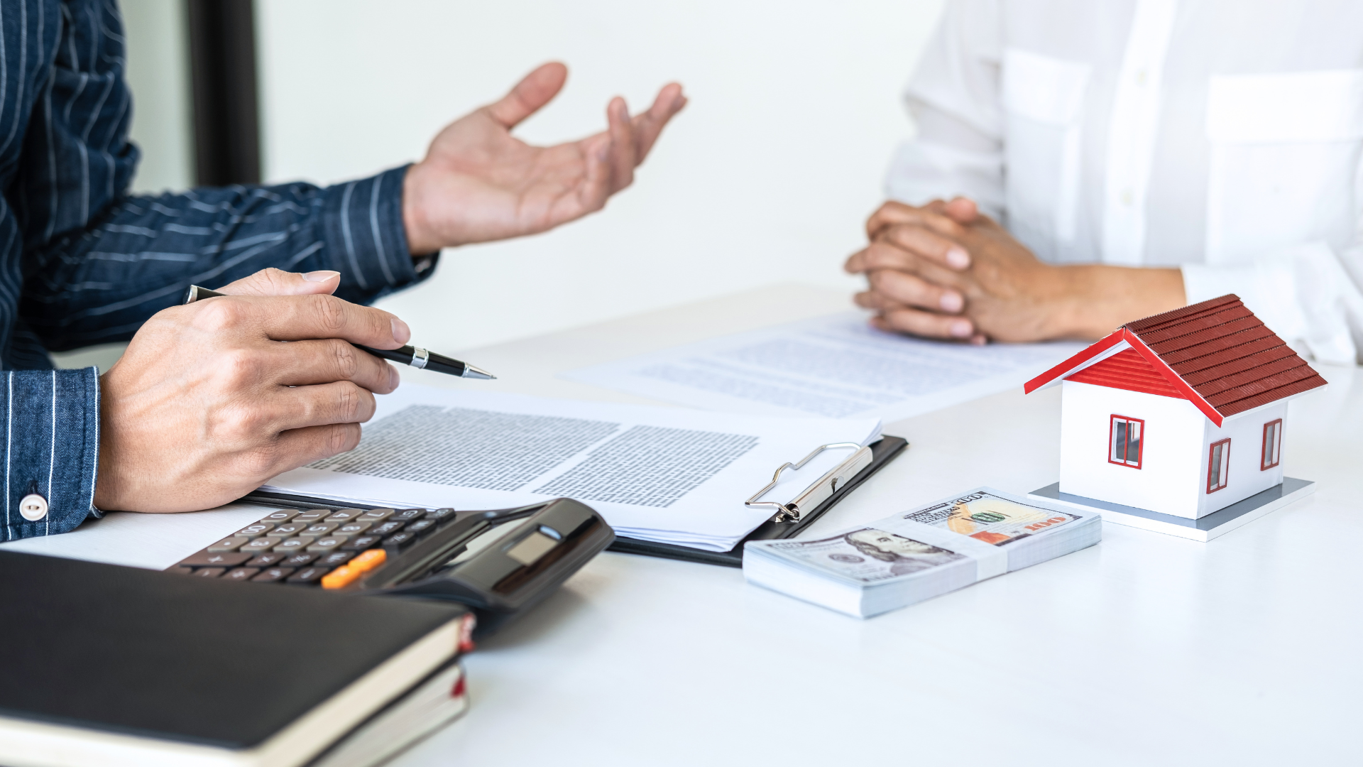 Person gesturing, discussing paperwork with another person at a desk with money, house model, and calculator.