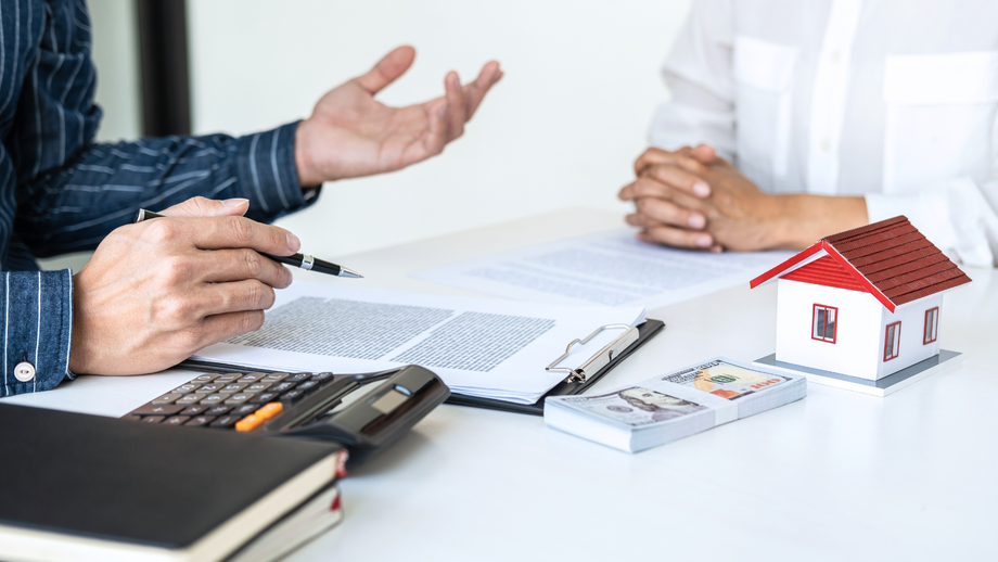 Person with pen gestures, discussing document with another person, near calculator, cash, and house model.