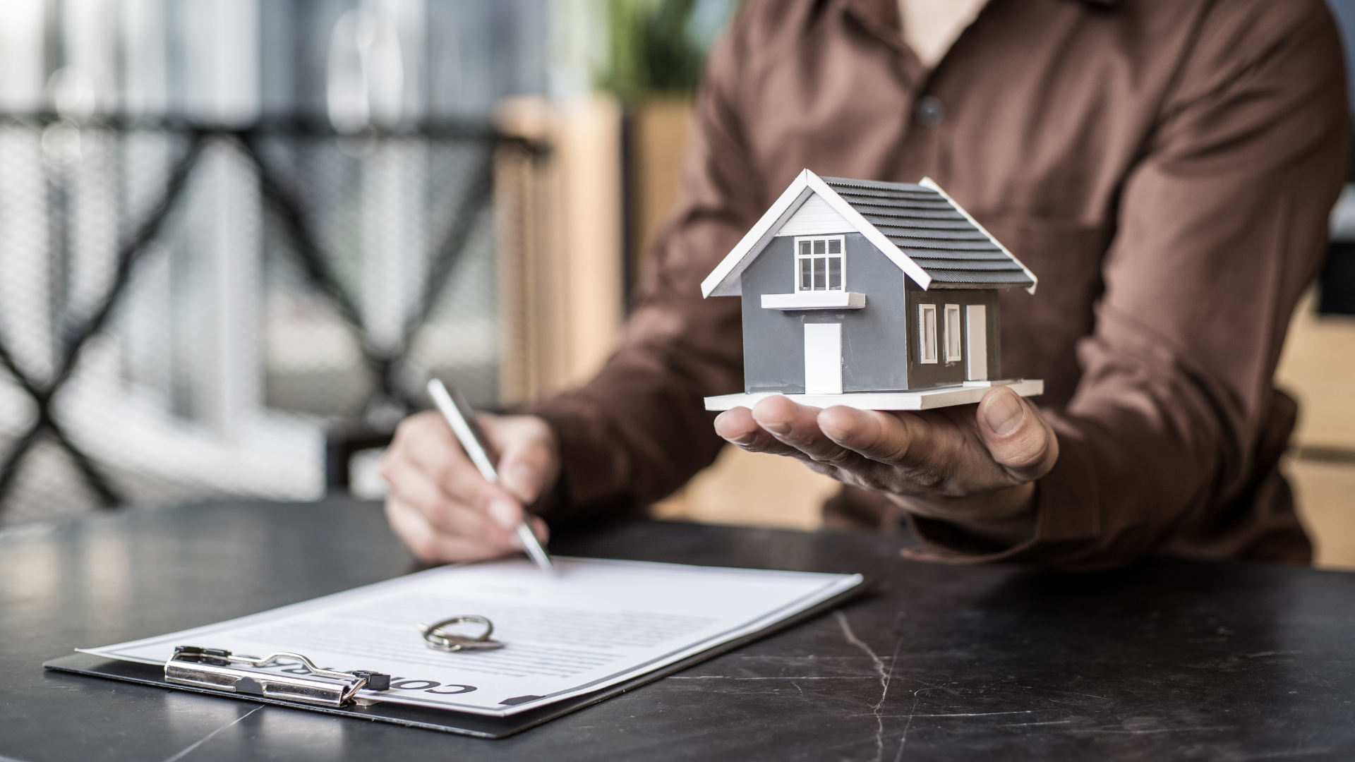 Person holding a miniature house, signing a document at a desk, symbolizing a real estate deal.