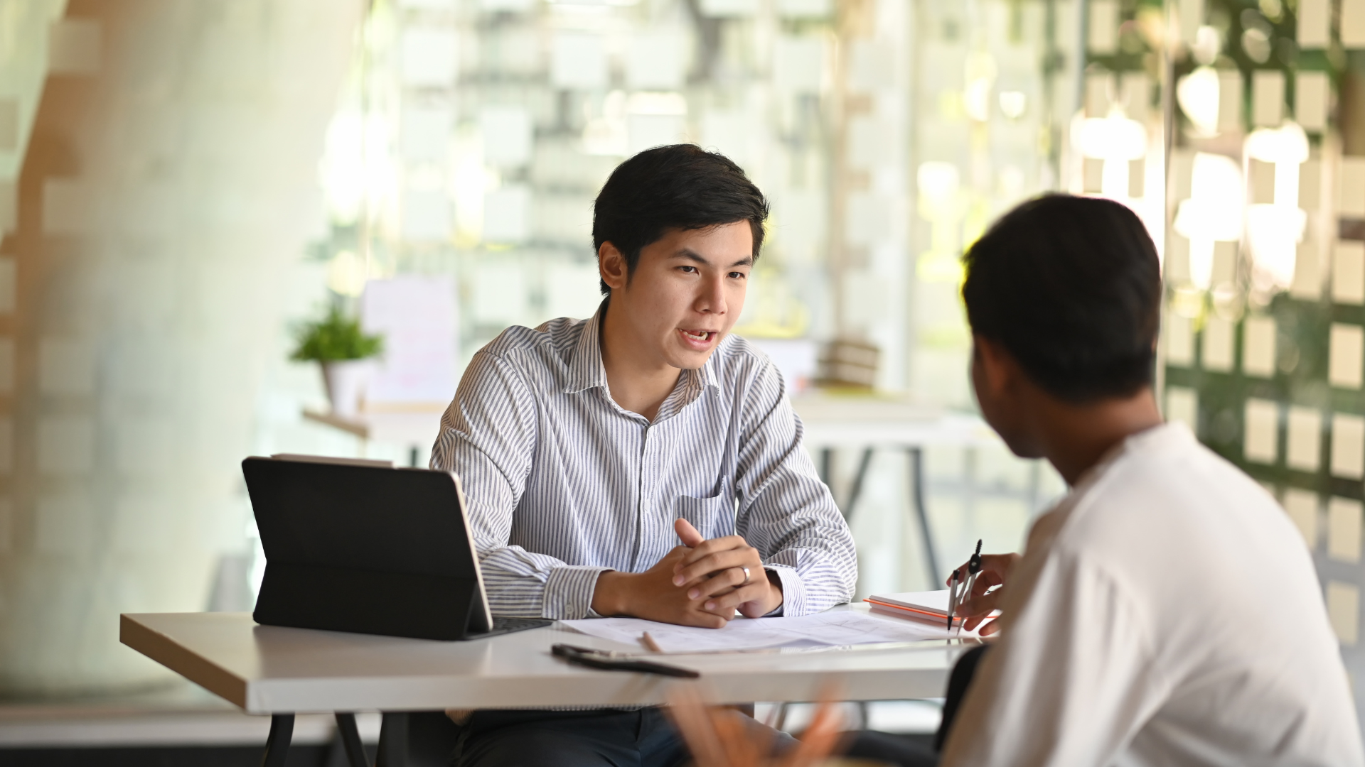 Two men at a table, one speaking, the other listening, in an office setting.