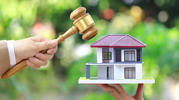 Hand holding a gavel over a miniature house, symbolizing a real estate auction.