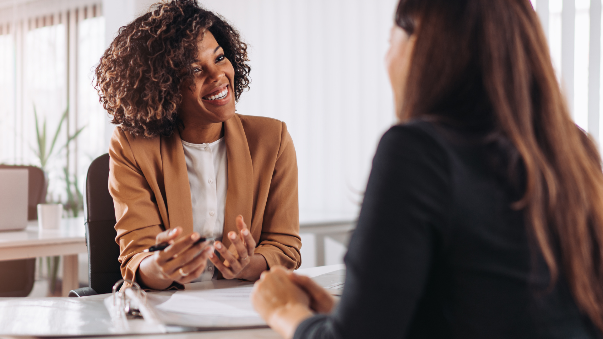 Woman in tan blazer smiles while speaking to another person at a desk.
