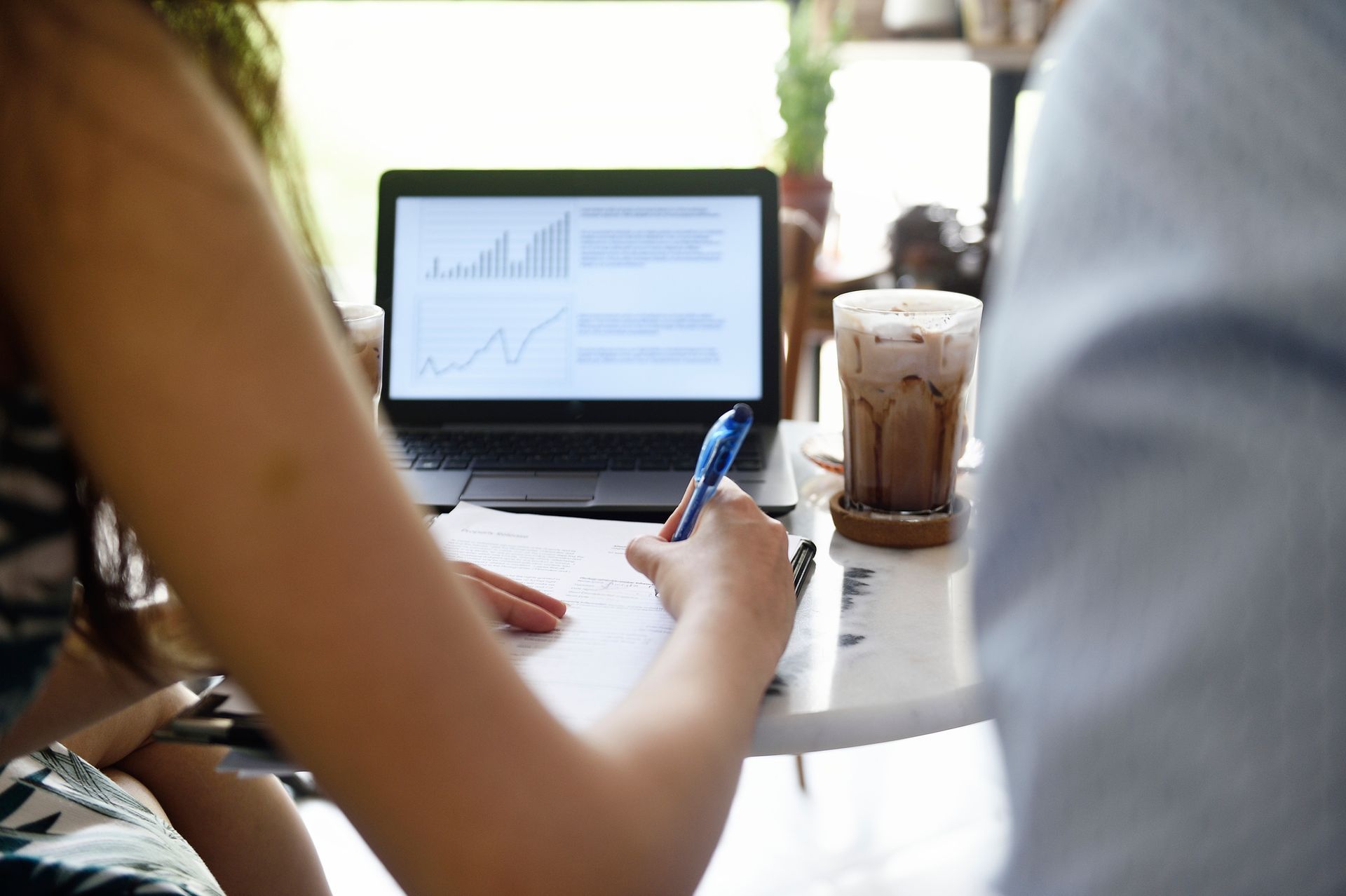 Woman writing on paper, working on laptop with charts, iced coffee on table.