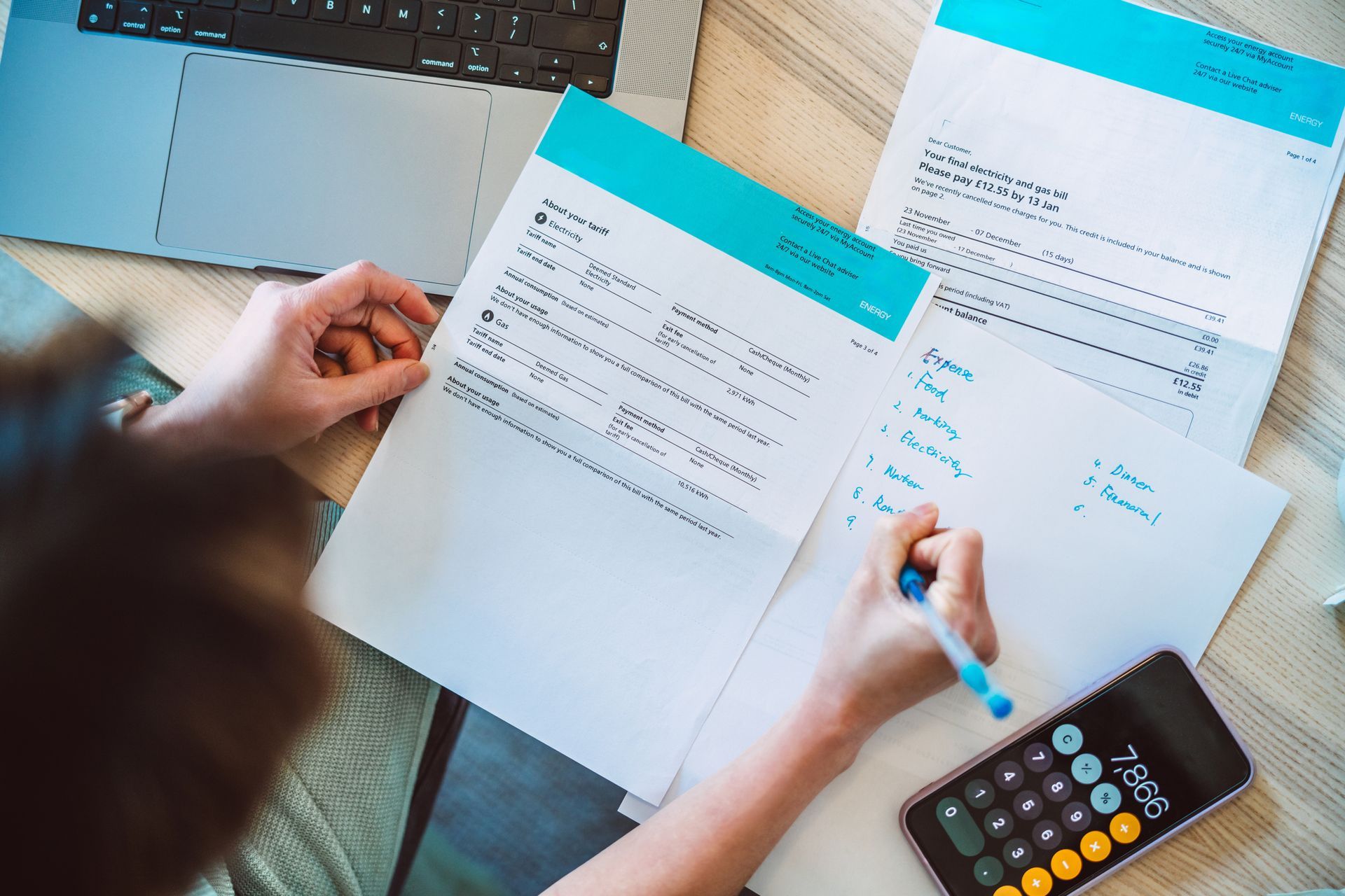 Person at a desk with papers, laptop, and calculator, writing notes.