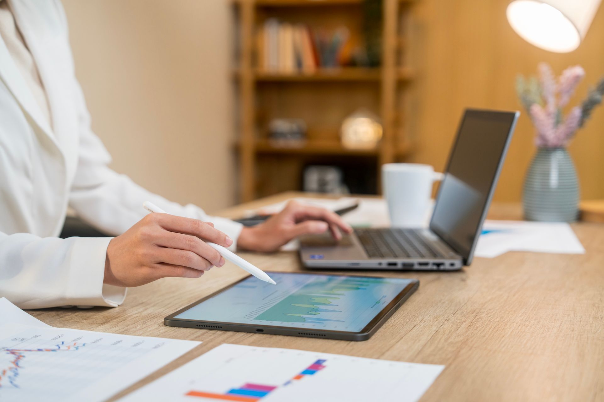 Woman using a tablet and laptop at a desk, analyzing data, home office setting.