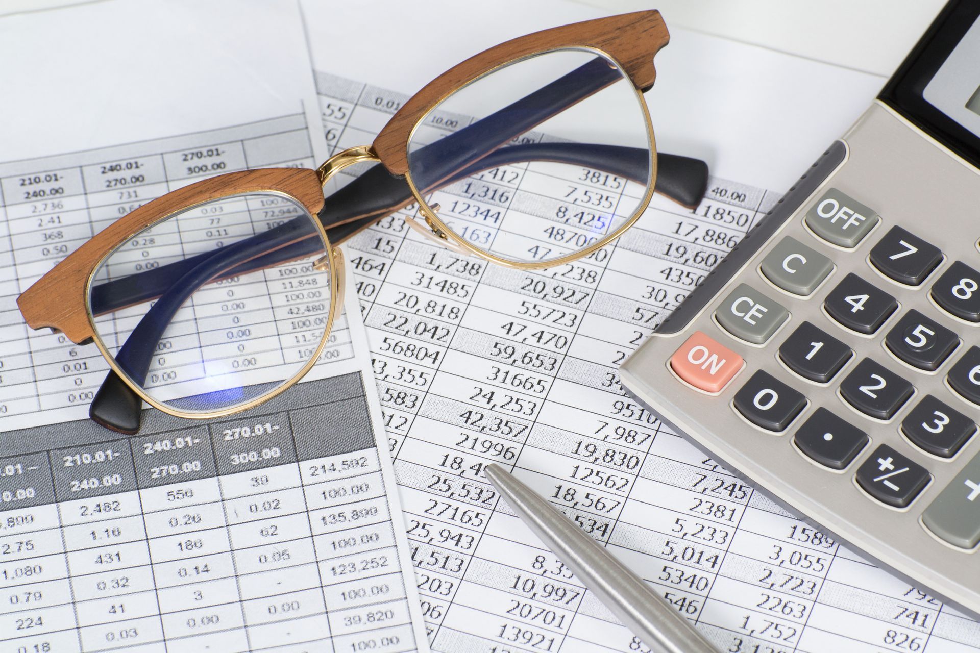 Glasses, calculator, and pen on a spreadsheet with numbers, implying financial work.