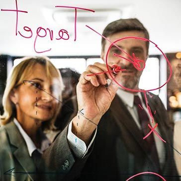 Two business people in suits, one drawing a circle with a red marker on a clear board.