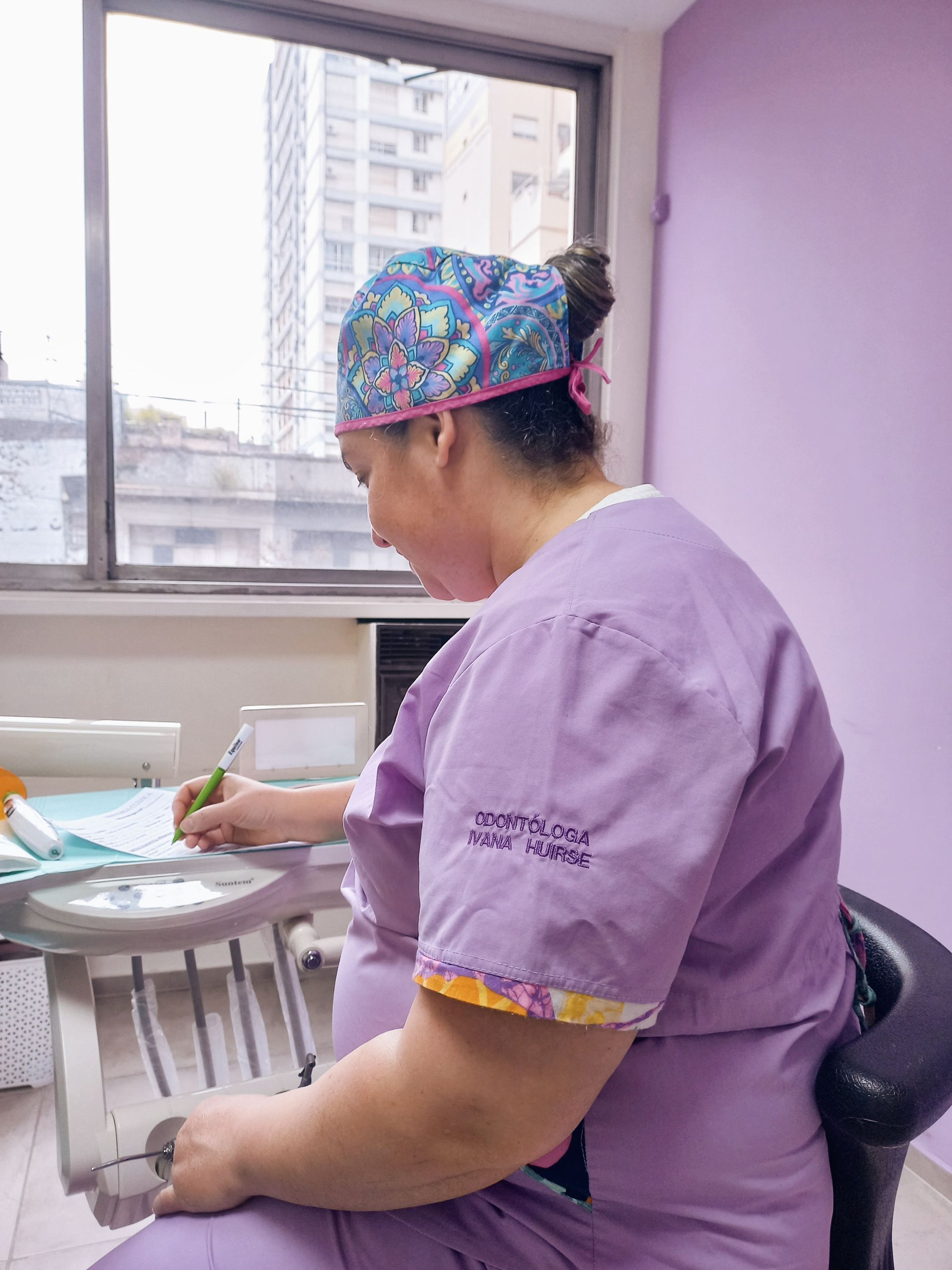 Una mujer con un uniforme morado está sentada en una silla dental escribiendo en un trozo de papel.