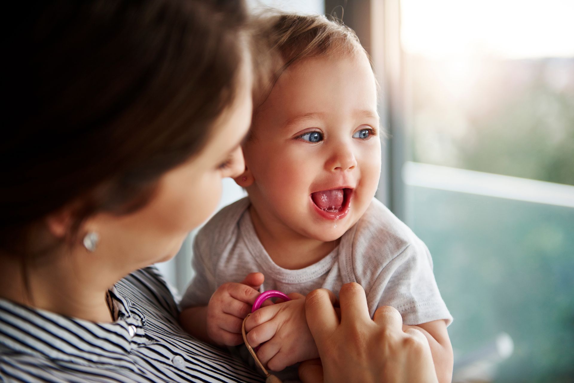 Woman holding a smiling baby near a window; baby with bright eyes and open mouth.